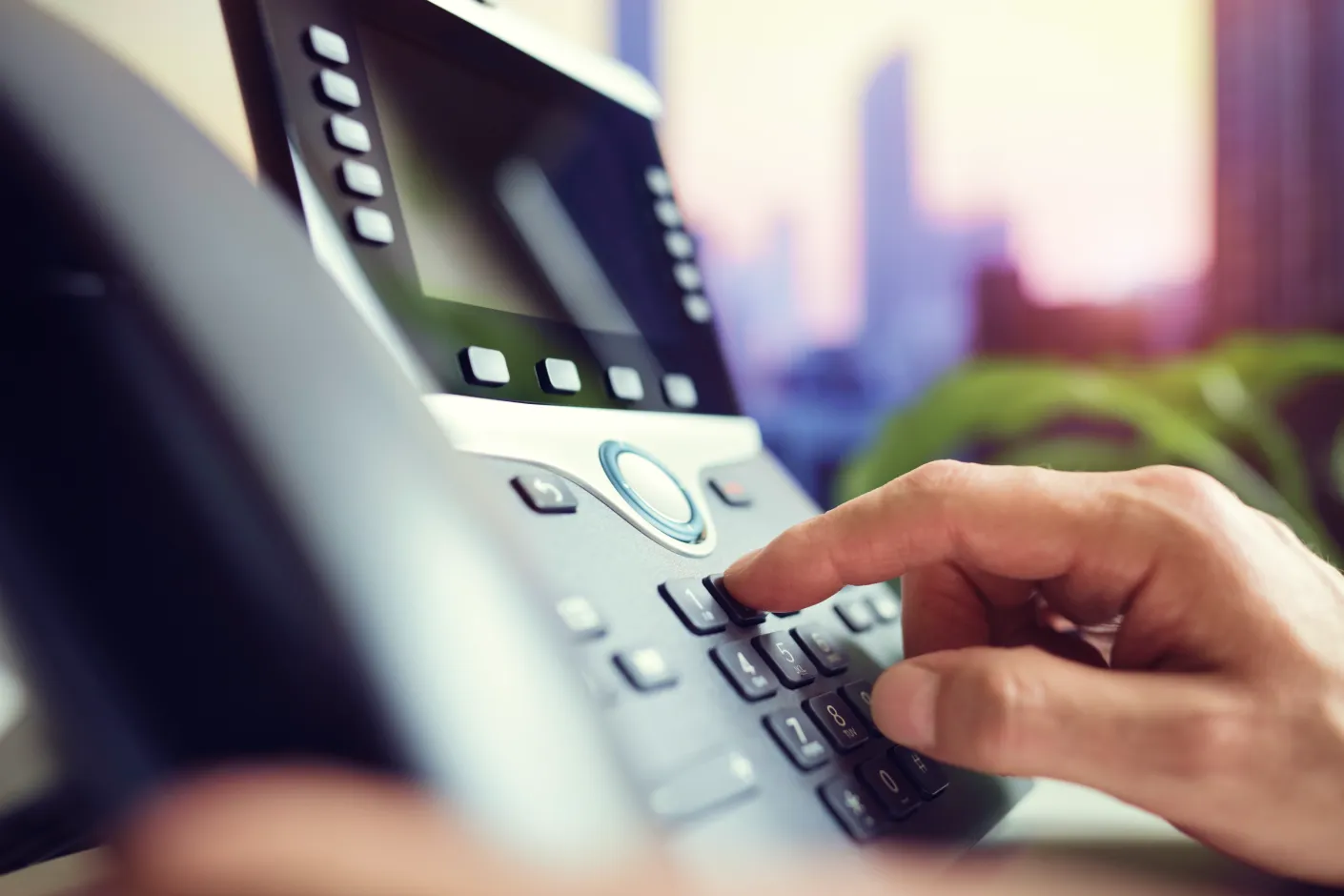 f-voip Close-up of a person dialing a number on an office desk phone, with blurred city buildings visible through the window in the background.