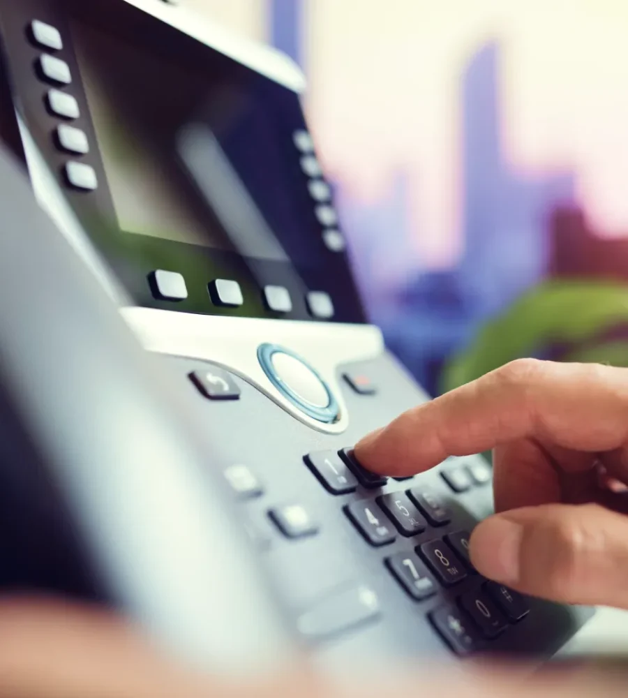 Close-up of a person dialing a number on an office desk phone, with blurred city buildings visible through the window in the background.